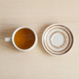 A white ceramic mug filled with amber-brown tea sits next to a matching saucer with concentric brown rings, set on a light wood surface.