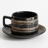 A black ceramic coffee cup with a square handle and matching saucer, decorated with horizontal metallic gold and bronze textured stripes, on a light gray background.