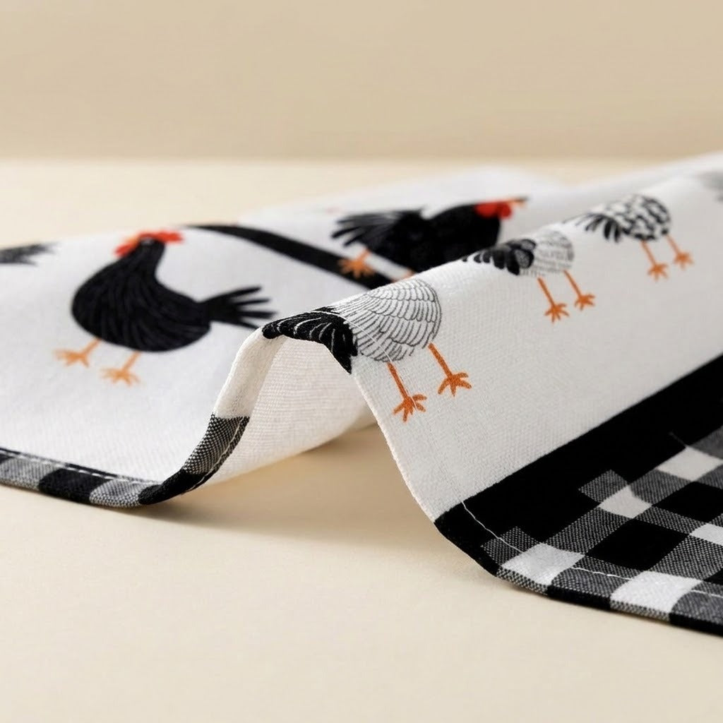 A folded white kitchen towel or napkin featuring a black, white, and gray chicken pattern with orange beaks and feet, and a black-and-white gingham check border, shown on a light beige background.