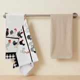 Two kitchen towels hanging from a metal rod on a beige wall. One towel is white with a black-and-white rooster pattern and a black-and-white gingham corner, while the other is a plain beige towel.