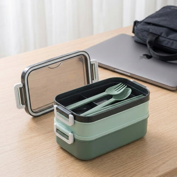 A two-tier bento lunch box in sage green and black, shown open with a matching green plastic utensil set inside. The lunch box is placed on a light wood desk next to a closed silver laptop and a black bag, with soft-focus curtains in the background.