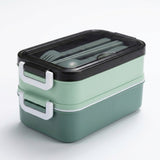 A two-tier bento lunch box with a mint green body, a darker green bottom layer, a black lid, and white handles, shown with gray utensils inside on a white background.