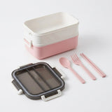 A two-tier bento box set in pastel pink and white, shown with its dark gray translucent lid and matching pink utensils (spoon, fork, chopsticks) on a clean white background.