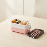 A pink and white two-compartment bento box filled with a colorful salad and toast, shown on a light wood table next to its black lid containing a matching pink utensil set.