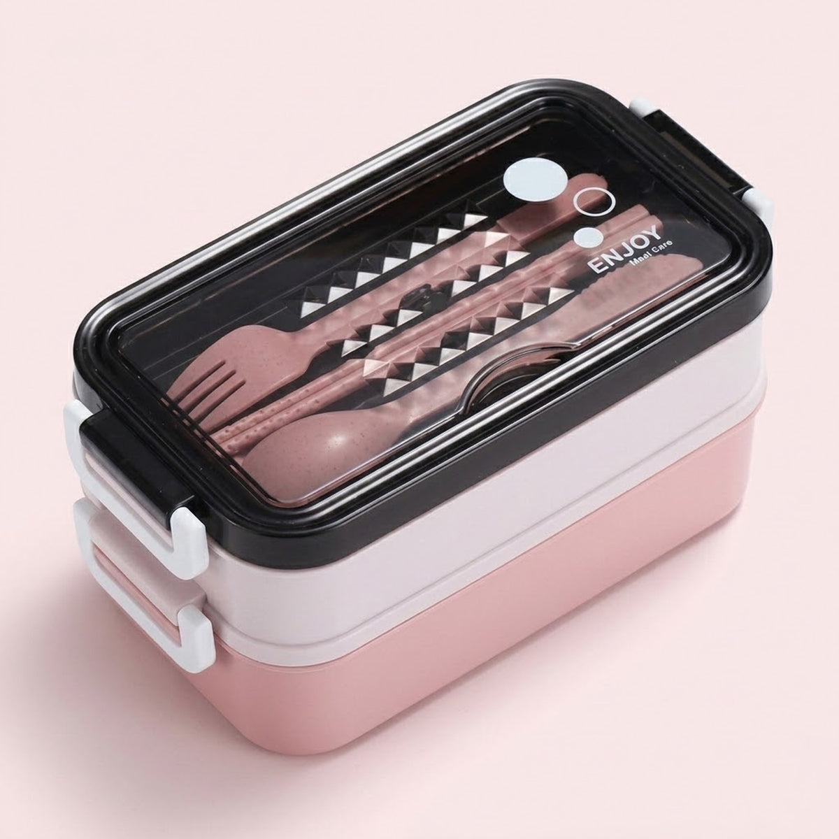 A two-tiered lunch box with a pink and white body, a black lid, and a clear window showing pink utensils inside, set against a solid pale pink background.