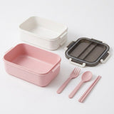 A set of minimalist bento containers and utensils on a clean white background. The set includes a pink container in the foreground, a white container in the back left, a dark gray translucent container in the back right, and matching pink utensils (fork, spoon, chopsticks) in the center.