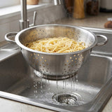 A stainless steel colander filled with cooked pasta, draining water into a sink in a kitchen setting.