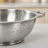 A stainless steel colander with two loop handles, shown on a light gray countertop with a blurred kitchen background.