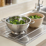 A stainless steel colander filled with broccoli sits on a sink drain board next to a beige bowl of broccoli in a kitchen setting.