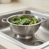 A stainless steel colander with side handles sits in a kitchen sink, draining green salad leaves.