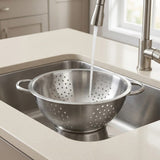 A stainless steel colander bowl with side handles sits in a kitchen sink under a running faucet, with a beige countertop and light cabinetry in the background.