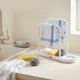 A white plastic double towel rack with a drain tray, shown on a bathroom or kitchen counter with two blue-striped towels, sponges, and a钢丝 ball. The background features a cream-colored wall, a sink faucet, and a soap dish.