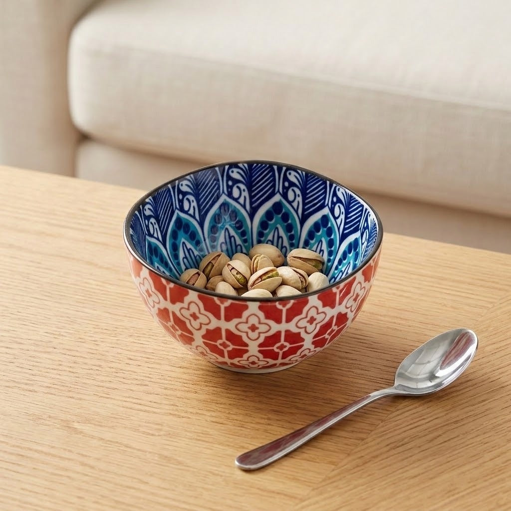 A decorative ceramic bowl filled with pistachios on a light wood table, with a silver spoon beside it and a blurred cream-colored sofa in the background.