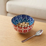 A decorative ceramic bowl filled with pistachios on a light wood table, with a silver spoon beside it and a blurred cream-colored sofa in the background.