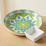 A decorative ceramic serving bowl with a green, blue, and white mandala pattern and a gold rim, shown on a light wood surface next to a small white square dish.