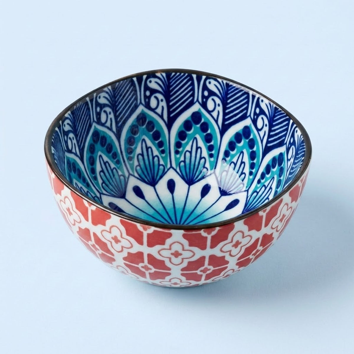 A decorative ceramic bowl with a red and white geometric pattern on the exterior and a blue and white floral mandala design on the interior, set against a light blue background.