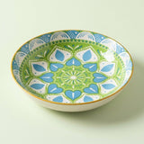A ceramic bowl with a colorful mandala-style pattern in green, blue, and white, featuring a gold rim, set against a solid pale green background.