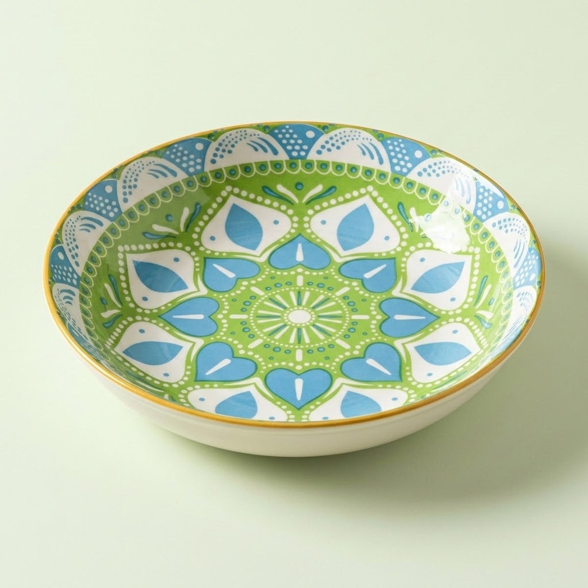 A ceramic bowl with a colorful mandala-style pattern in green, blue, and white, featuring a gold rim, set against a solid pale green background.