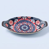 A decorative ceramic serving dish with a hand-painted mandala pattern in navy blue, red, white, and black, featuring dark brown handles on a light blue background.
