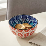 A ceramic bowl with a red and white geometric pattern on the outside and a blue and white floral pattern on the inside, filled with crackers on a white table next to a beige napkin.