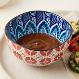 A decorative ceramic bowl with a red and white geometric pattern on the outside and a blue and white leaf pattern on the inside, filled with a brown sauce, set on a light beige plate with grilled vegetables.