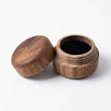 A small round wooden jar with its lid off, showing a black interior, photographed on a clean white background.