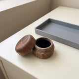 A small round wooden jar with its lid off, placed on a light cream tabletop next to a rectangular gray tray.