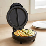 A black round electric waffle maker or omelet maker sits open on a light wood countertop, cooking two folded omelets with visible green herbs and red pepper pieces. A stack of white plates and a tiled kitchen wall are visible in the background.
