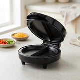 A black round sandwich maker or waffle press shown open on a light countertop, with bowls of chopped vegetables and shredded cheese blurred in the background.