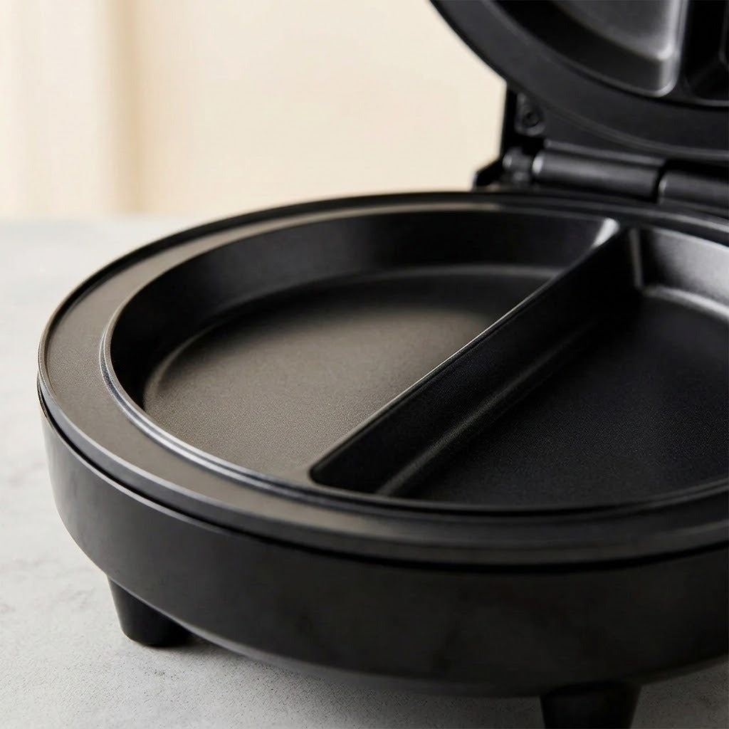 Close-up of a black round sandwich maker or waffle maker with the lid open, showing a divided cooking plate on a light gray surface.
