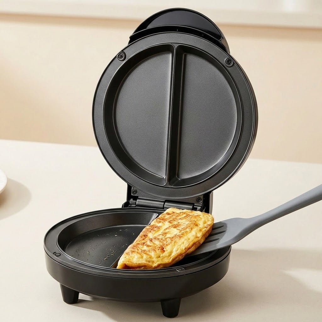 A black electric mini waffle maker with its lid open, showing a golden-brown omelet being lifted by a gray spatula on a light beige countertop.