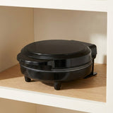 A black round waffle maker sitting on a wooden shelf inside a white cabinet or wall niche.