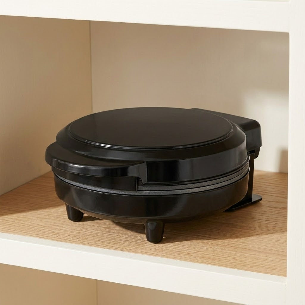 A black round waffle maker sitting on a wooden shelf inside a white cabinet or wall niche.