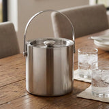 A brushed stainless steel ice bucket with a top handle and lid, set on a wooden dining table next to two clear drinking glasses with ice.