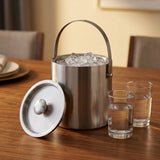 A stainless steel ice bucket with a handle, filled with ice, sits on a wooden table with its lid placed beside it and two clear drinking glasses nearby. The setting appears to be a dining or kitchen environment with softly blurred chairs and tableware in the background.