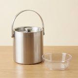 A stainless steel ice bucket with a carrying handle and lid, shown next to a clear plastic bowl on a light wood surface against a beige background.