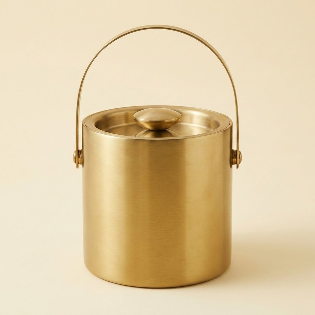 A brushed brass metal ice bucket with a matching lid and a curved metal carrying handle, shown on a solid light beige background.