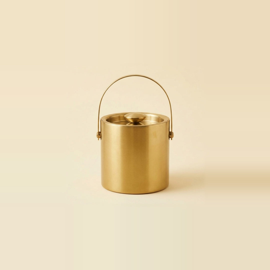 A brushed brass cylindrical ice bucket with a matching lid and a curved metal handle, centered against a solid cream background.
