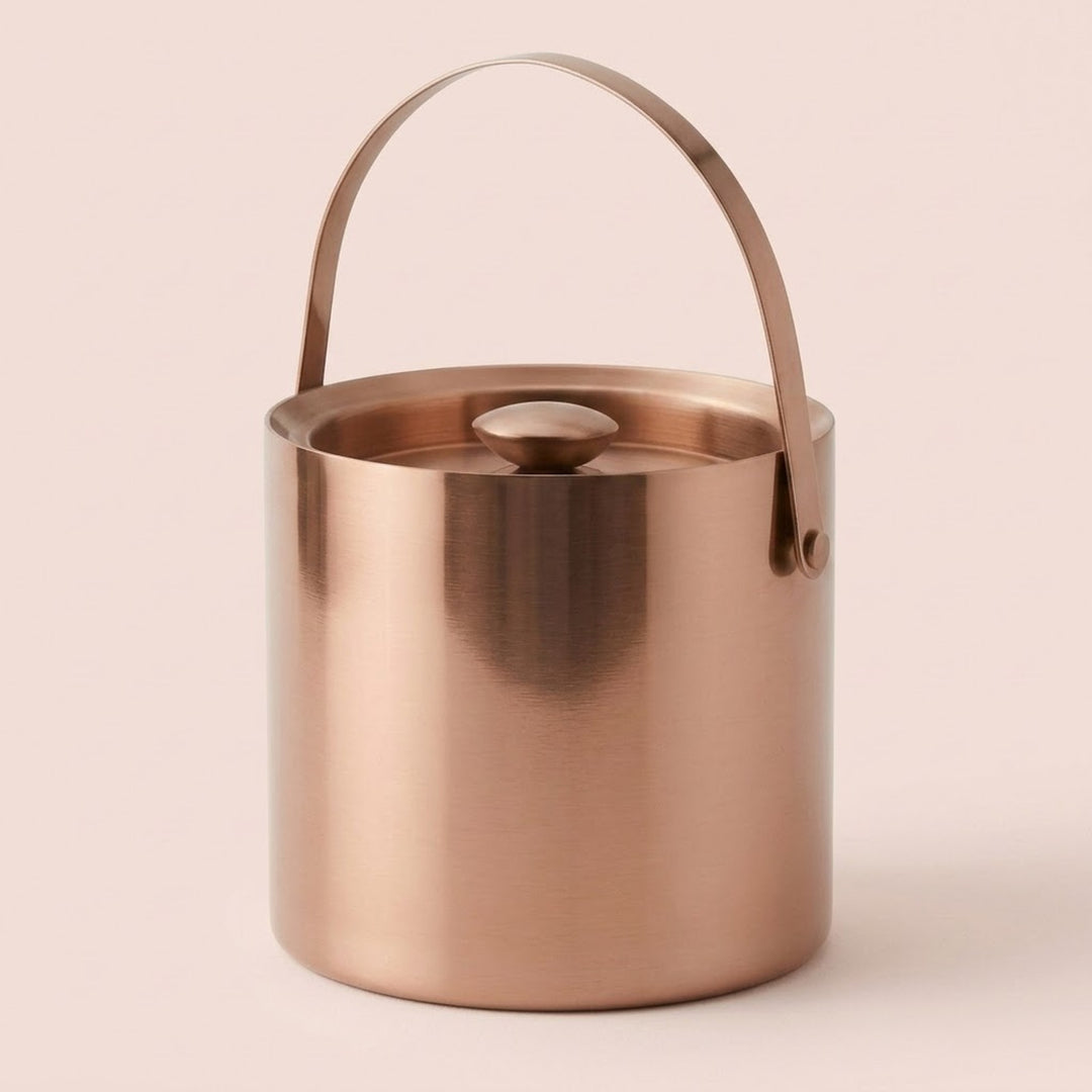 A brushed copper ice bucket with a matching metal handle and lid, shown on a solid pale pink background.