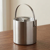 A brushed stainless steel cylindrical container with a matching lid and a curved metal handle, shown on a wooden tabletop against a soft beige indoor background.