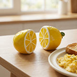 Two ceramic lemon-shaped salt and pepper shakers sit on a light wood table next to a plate of scrambled eggs and toast, with a softly blurred kitchen window in the background.