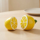 Two yellow lemon-shaped ceramic objects, one whole and one cut in half to show the cross-section, placed on a light wooden tabletop with a blurred plate in the background.