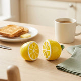 Two ceramic lemon-shaped salt and pepper shakers on a light wood table in a breakfast setting, with toast, butter, a mug of coffee, and a napkin nearby.