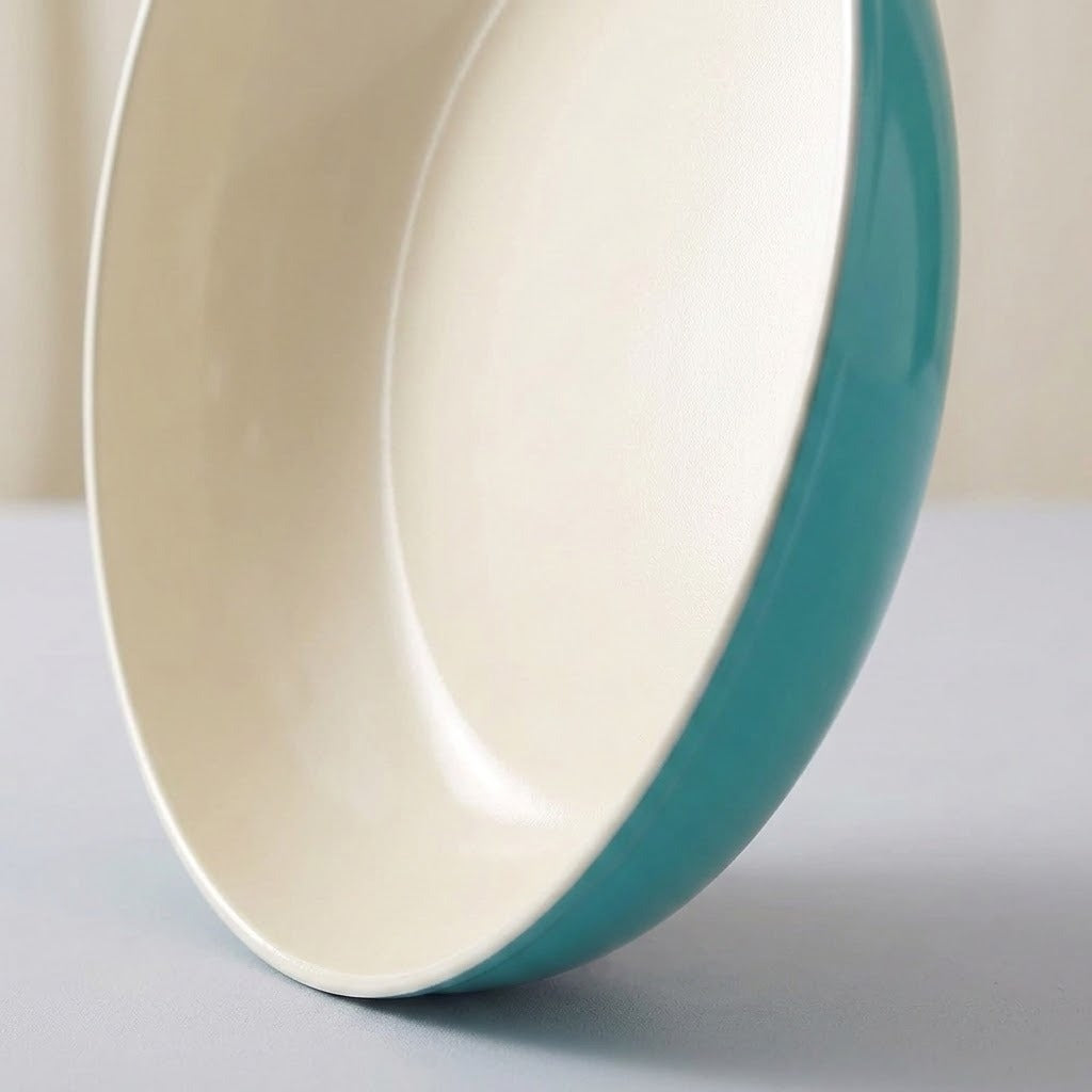 A close-up of a shallow round bowl or plate with a glossy off-white interior and a smooth teal exterior, photographed on a light neutral surface.