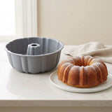 A gray fluted bundt cake pan sits on a white marble countertop next to a golden-brown bundt cake on a white plate, with a beige cloth and a softly lit window in the background.