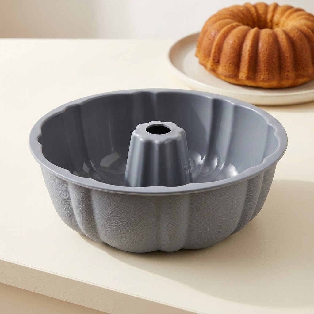 A gray fluted bundt cake pan sits on a light beige tabletop, with a baked ring cake on a plate blurred in the background.