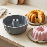 A gray fluted bundt cake mold sits on a light beige countertop, with two baked bundt cakes on plates in the background—one golden yellow and one pink—suggesting use of the mold.