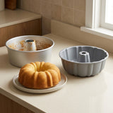 A fluted bundt cake pan in light gray sits on a beige countertop next to a baked golden-brown ring cake on a small plate, with an empty metal bundt pan in the background.