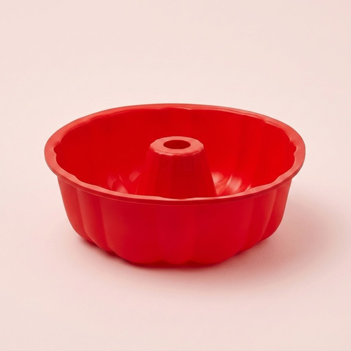 A bright red fluted bundt cake pan with a central hole, photographed on a clean, solid, pale pinkish-beige background.