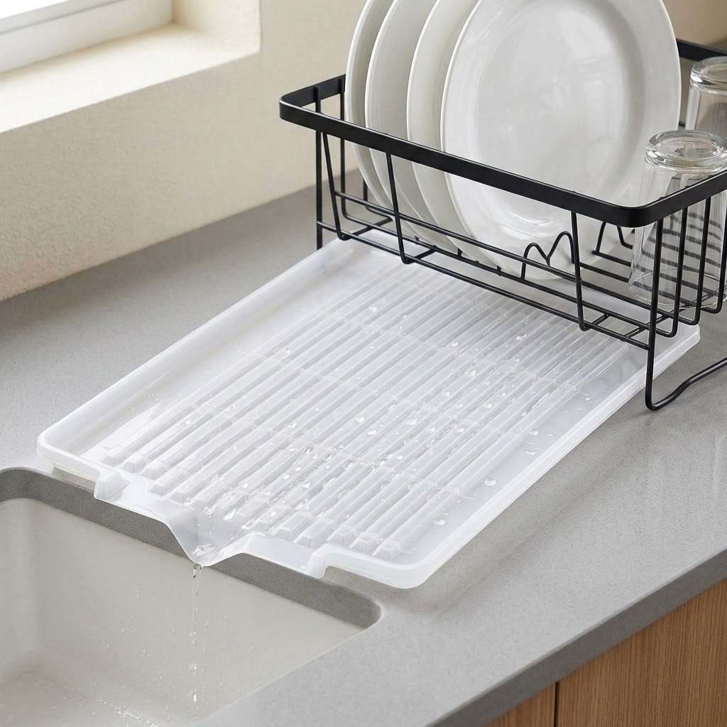 A white plastic under-sink drying rack with slotted drain holes sits on a gray kitchen countertop next to a sink, with a black wire dish rack holding white plates in the background.